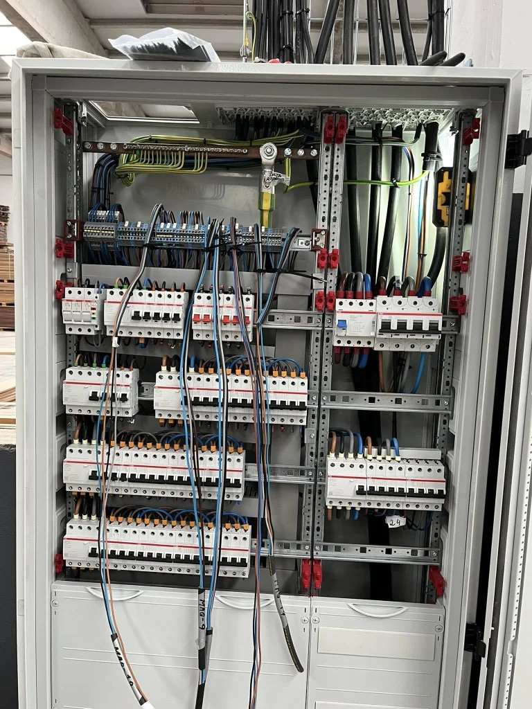 Transformador cinza com aletas de resfriamento instalado em compartimento metálico aberto. Cabos grossos pretos, vermelhos e amarelos estão fixados nos terminais com abraçadeiras. Etiquetas de advertência elétrica aparecem na parte frontal. O compartimento possui ventilação superior, reforçando ambiente técnico de distribuição de energia.